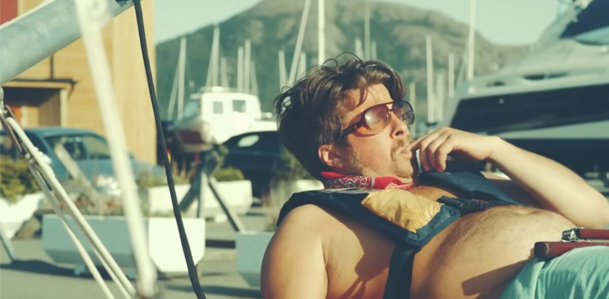 Man met Ray-Ban zonnebril, Spinlock reddingsvest (ISO gecertificeerd), nippend aan koffie op jachthaven; boten, bergen, zon.